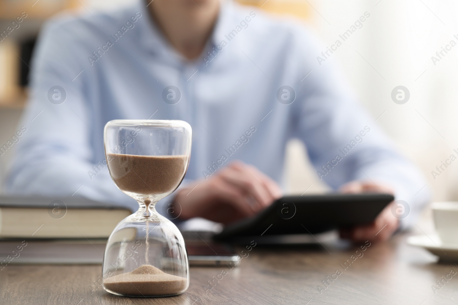 Photo of Hourglass with flowing sand on desk. Man using calculator indoors, selective focus