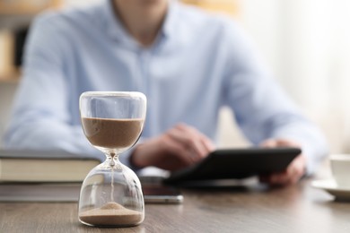 Photo of Hourglass with flowing sand on desk. Man using calculator indoors, selective focus