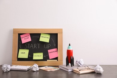 Photo of Blackboard with words Start Up, stationery and toy rocket on grey table, space for text