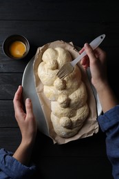 Photo of Woman spreading egg yolk onto raw braided bread at black wooden table, top view. Traditional Shabbat challah