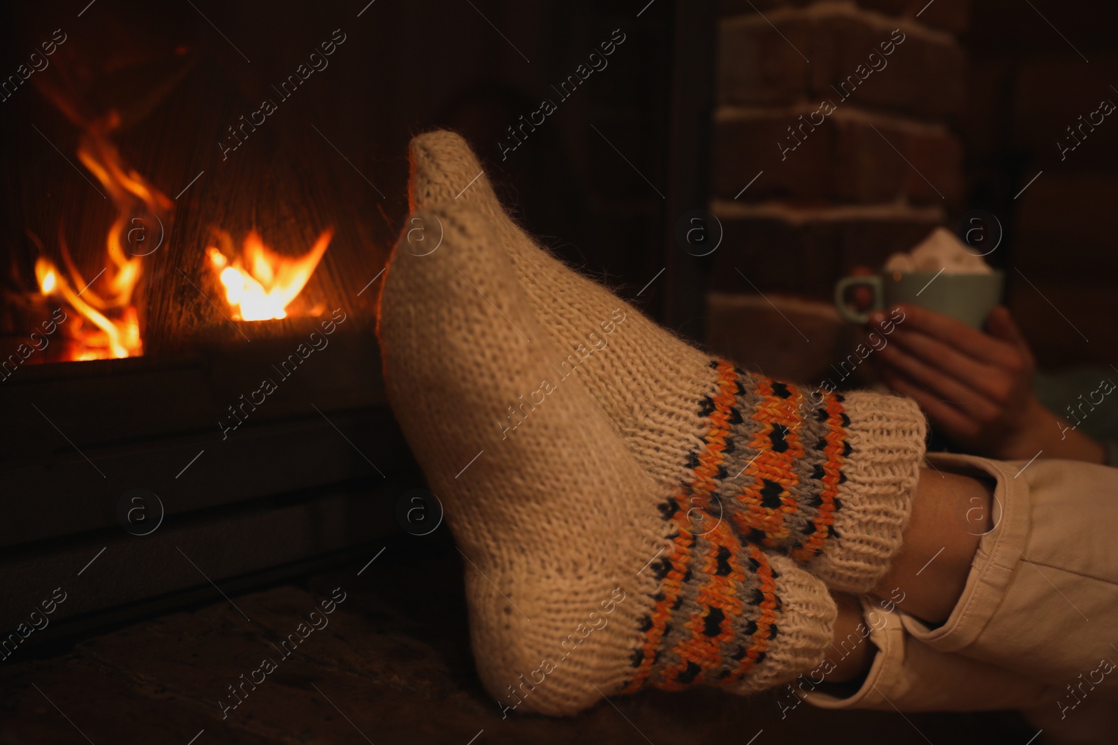 Photo of Woman in knitted socks with cup of sweet cocoa near fireplace indoors, closeup