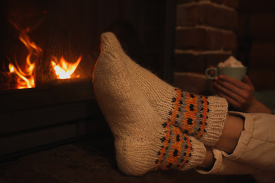 Photo of Woman in knitted socks with cup of sweet cocoa near fireplace indoors, closeup