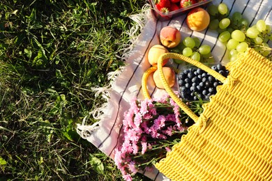 Yellow wicker bag with beautiful flowers, fruits and berries on picnic blanket outdoors, top view