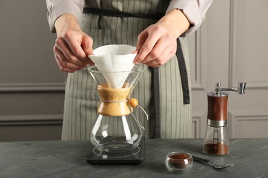 Man putting paper filter into glass chemex coffeemaker at gray table, closeup