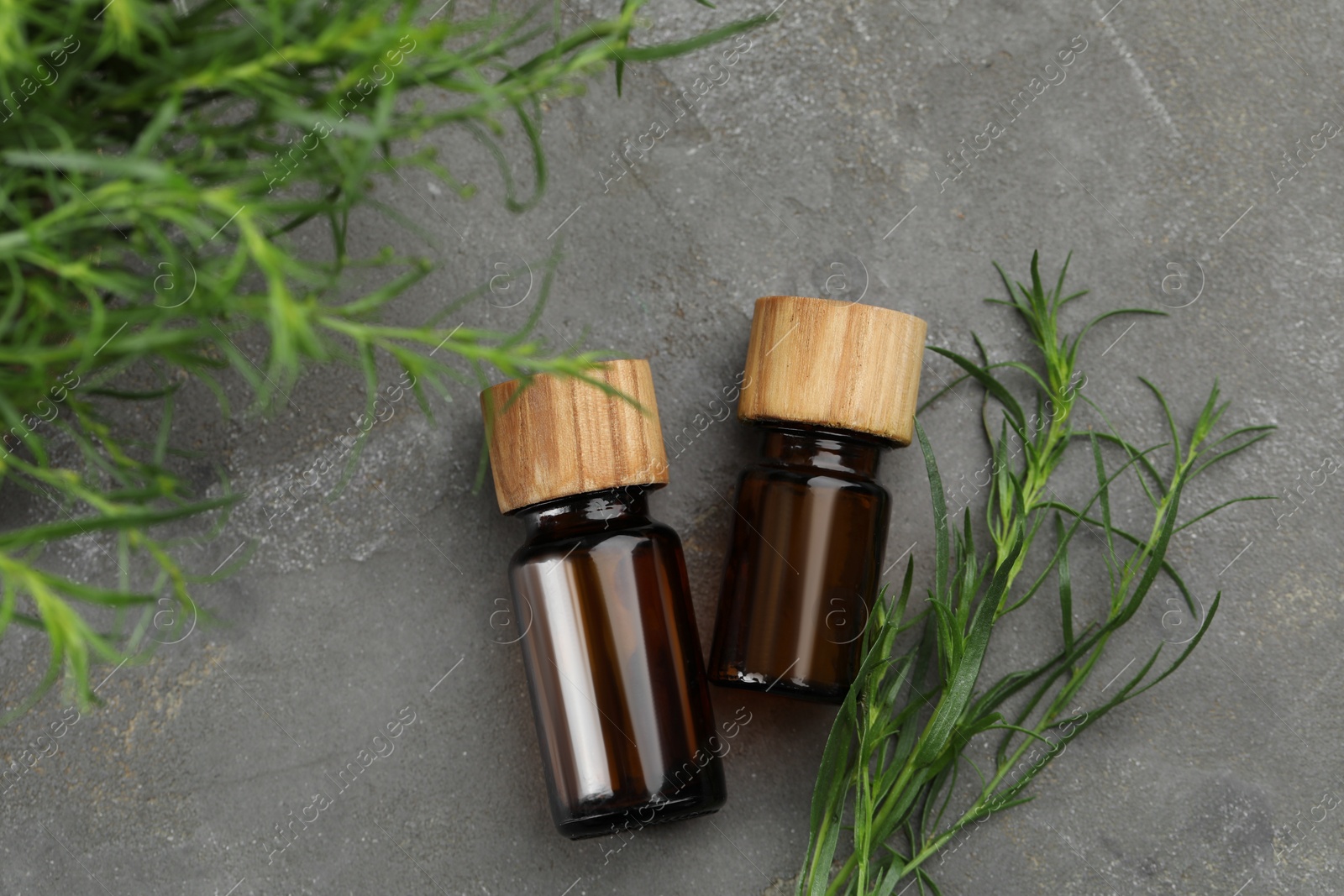 Photo of Bottles of essential oil and fresh tarragon leaves on grey table, flat lay