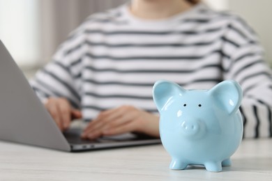 Photo of Financial savings. Woman using laptop at white wooden table indoors, focus on piggy bank