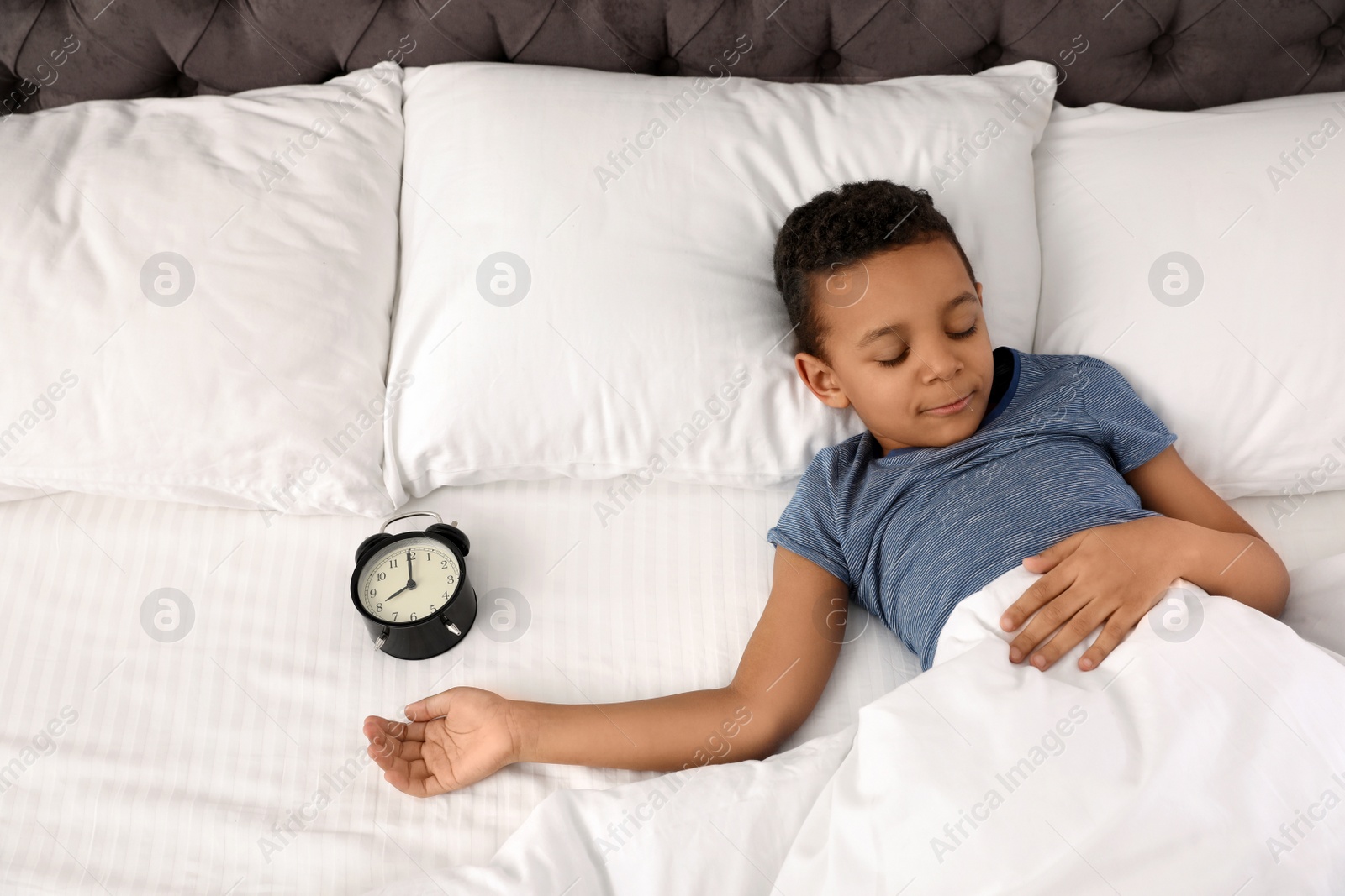 Photo of Cute little African-American boy with alarm clock sleeping in bed