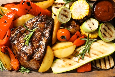 Photo of Delicious barbecued steak served with garnish and sauce on wooden board, top view