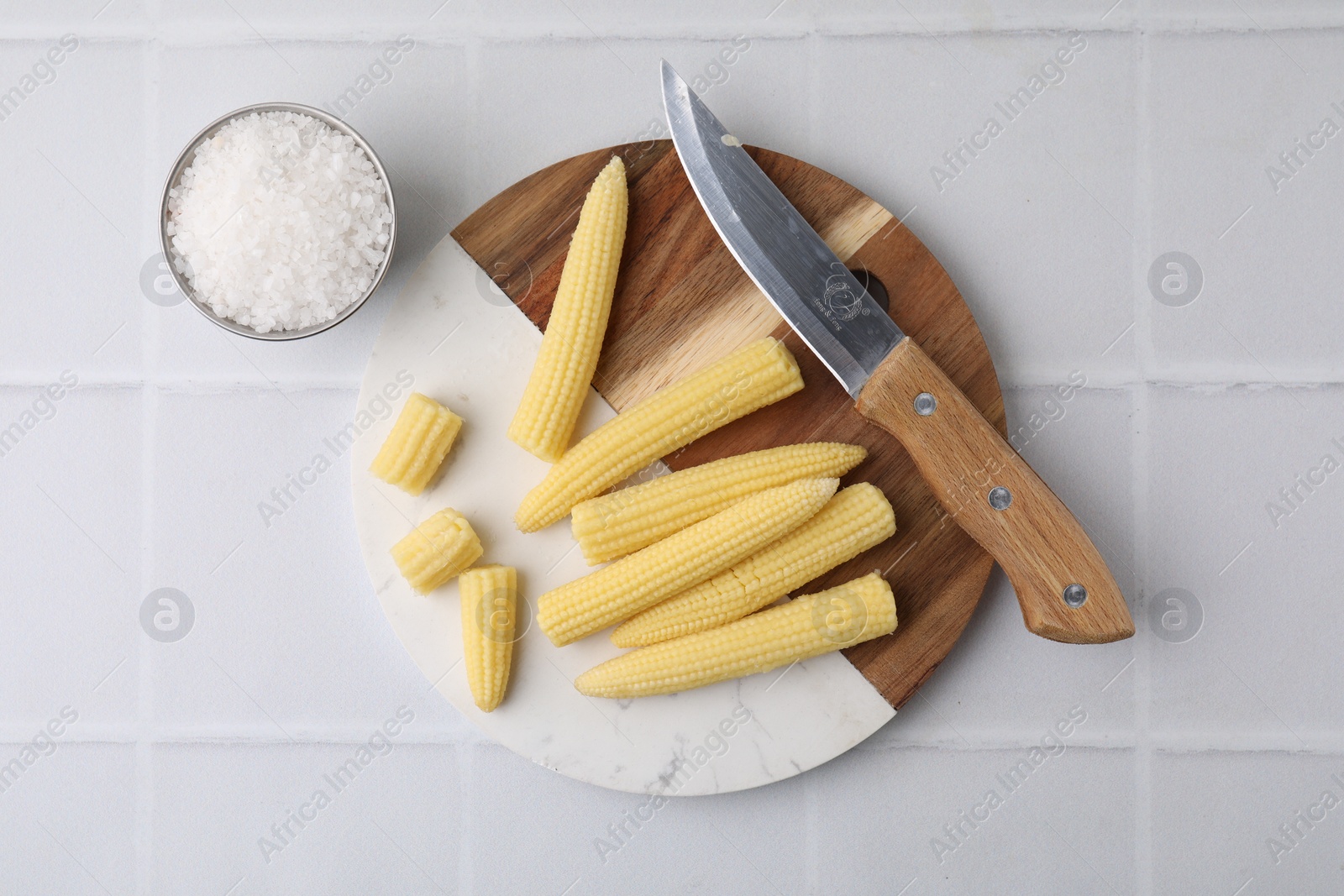 Photo of Tasty fresh yellow baby corns and knife on white tiled table, top view