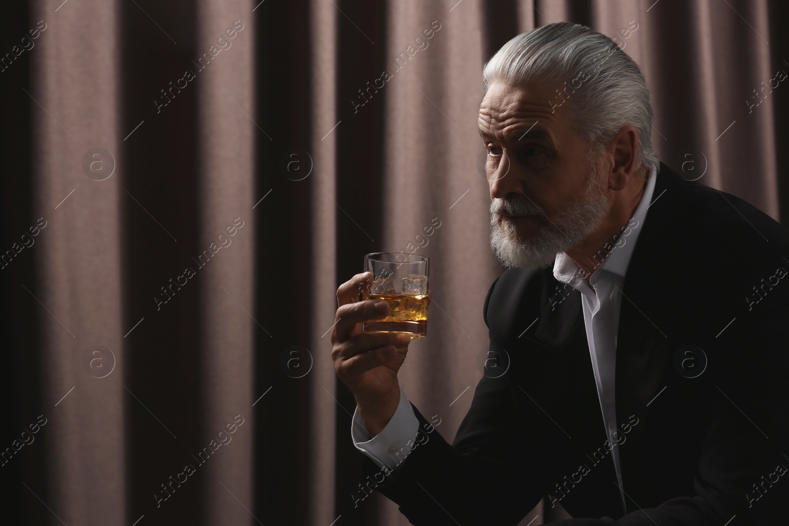 Photo of Senior man in suit holding glass of whiskey with ice cubes on brown background. Space for text