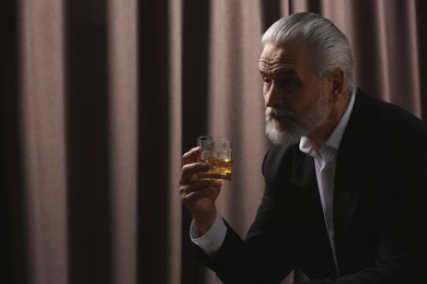 Photo of Senior man in suit holding glass of whiskey with ice cubes on brown background. Space for text