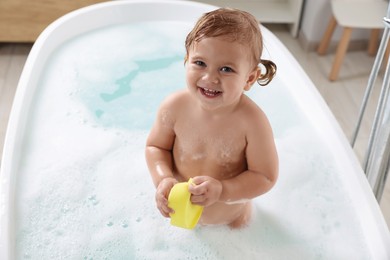 Photo of Cute little girl with sponge taking foamy bath at home