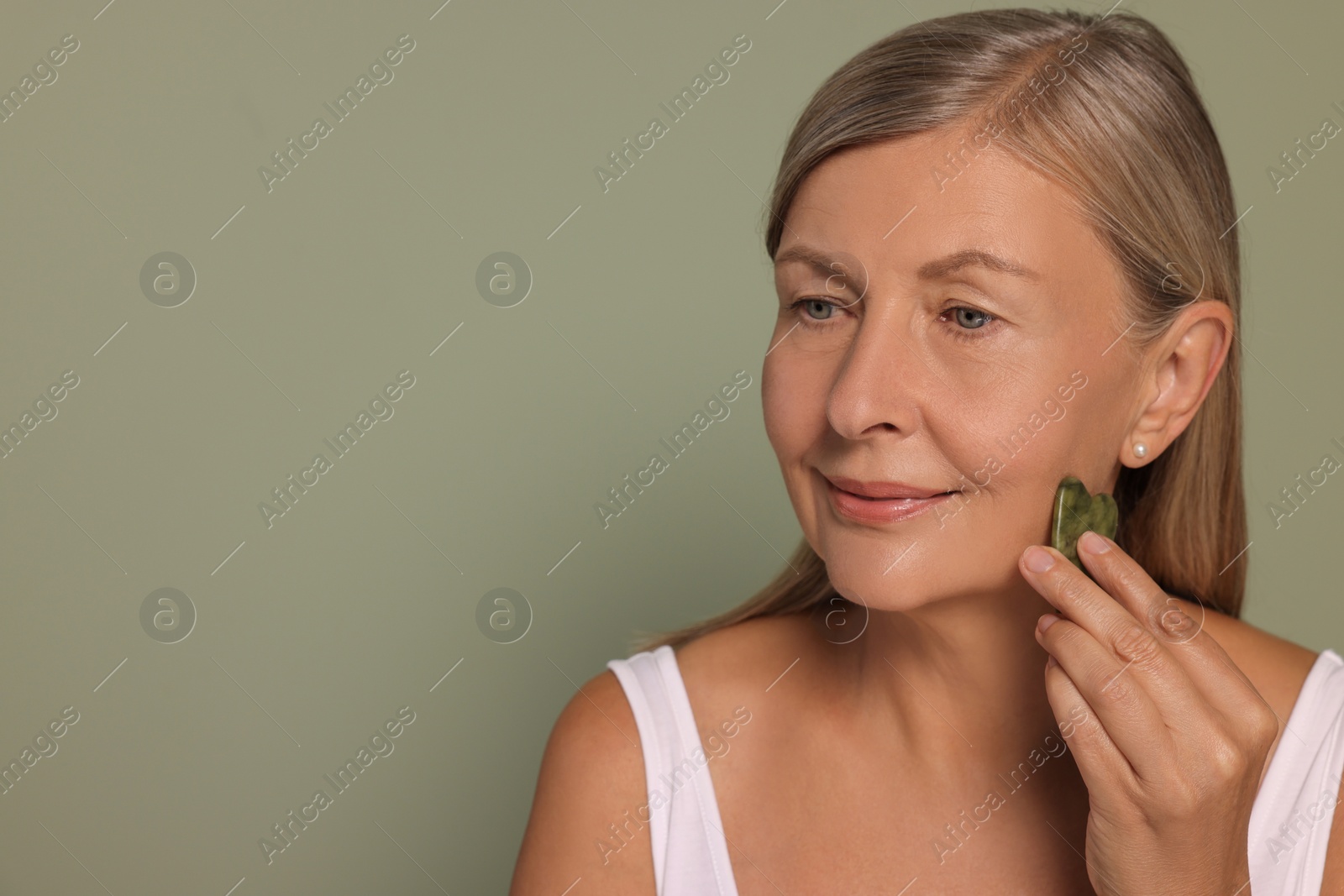 Photo of Woman massaging her face with jade gua sha tool on green background. Space for text