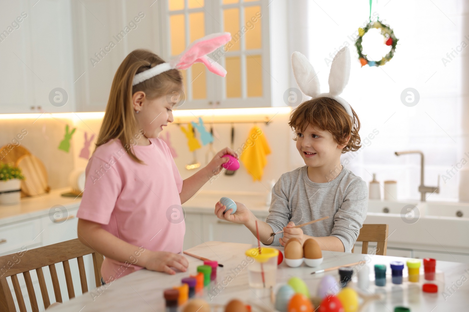 Photo of Easter celebration. Cute children with bunny ears having fun while painting eggs at white marble table in kitchen