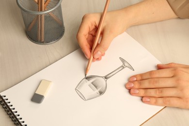 Photo of Woman drawing glass of wine with graphite pencil at light wooden table, closeup