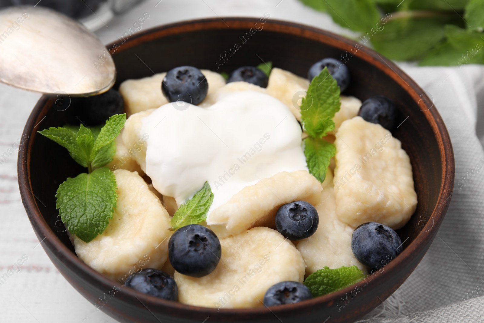 Photo of Bowl of tasty lazy dumplings with blueberries, sour cream and mint leaves on white wooden table, closeup