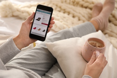 Photo of Woman with cup of coffee using smartphone for online shopping indoors, closeup