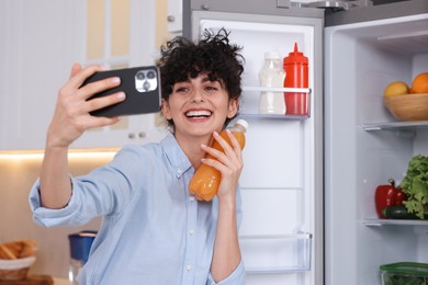 Smiling food blogger with bottle of juice taking selfie near fridge in kitchen