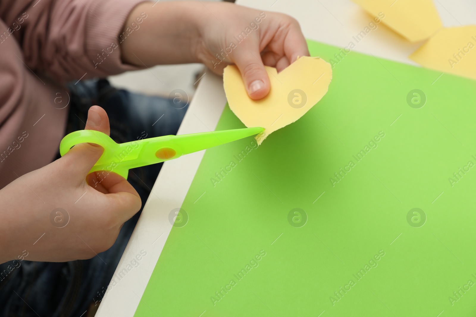 Photo of Child cutting out paper heart with plastic scissors at table, closeup. Space for text