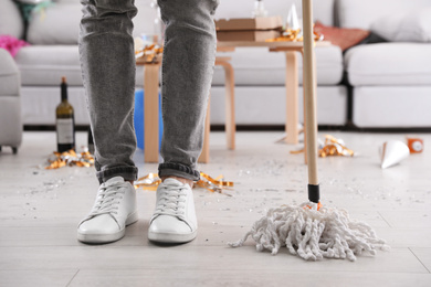 Photo of Young man with mop cleaning messy room after party, closeup of legs