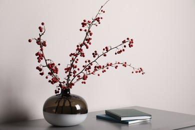 Hawthorn branches with red berries in vase and books on grey table indoors