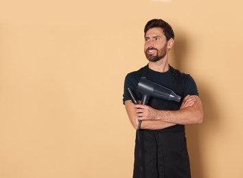 Photo of Smiling hairdresser in apron holding dryer and brush on light brown background, space for text