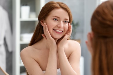 Photo of Smiling woman with freckles and cream on her face near mirror in bathroom