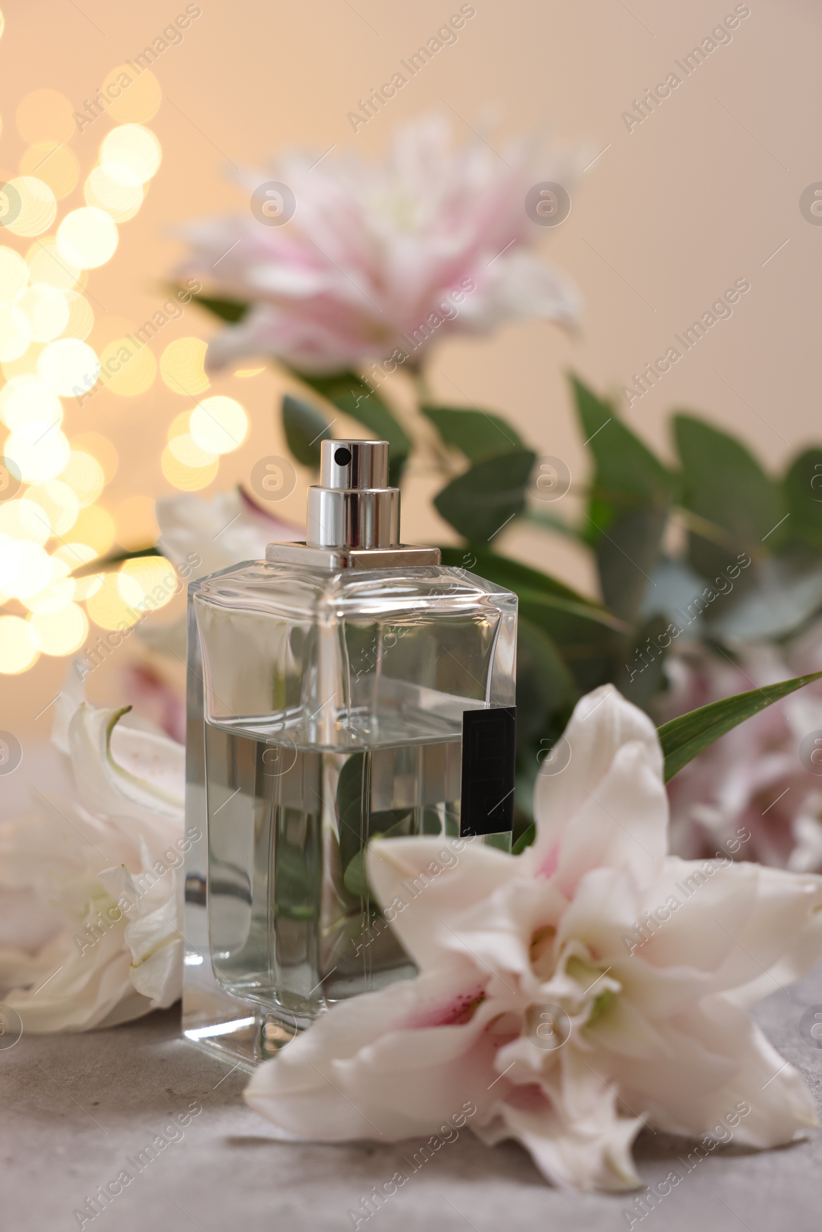 Photo of Bottle of perfume and beautiful lily flowers on table against beige background with blurred lights, closeup