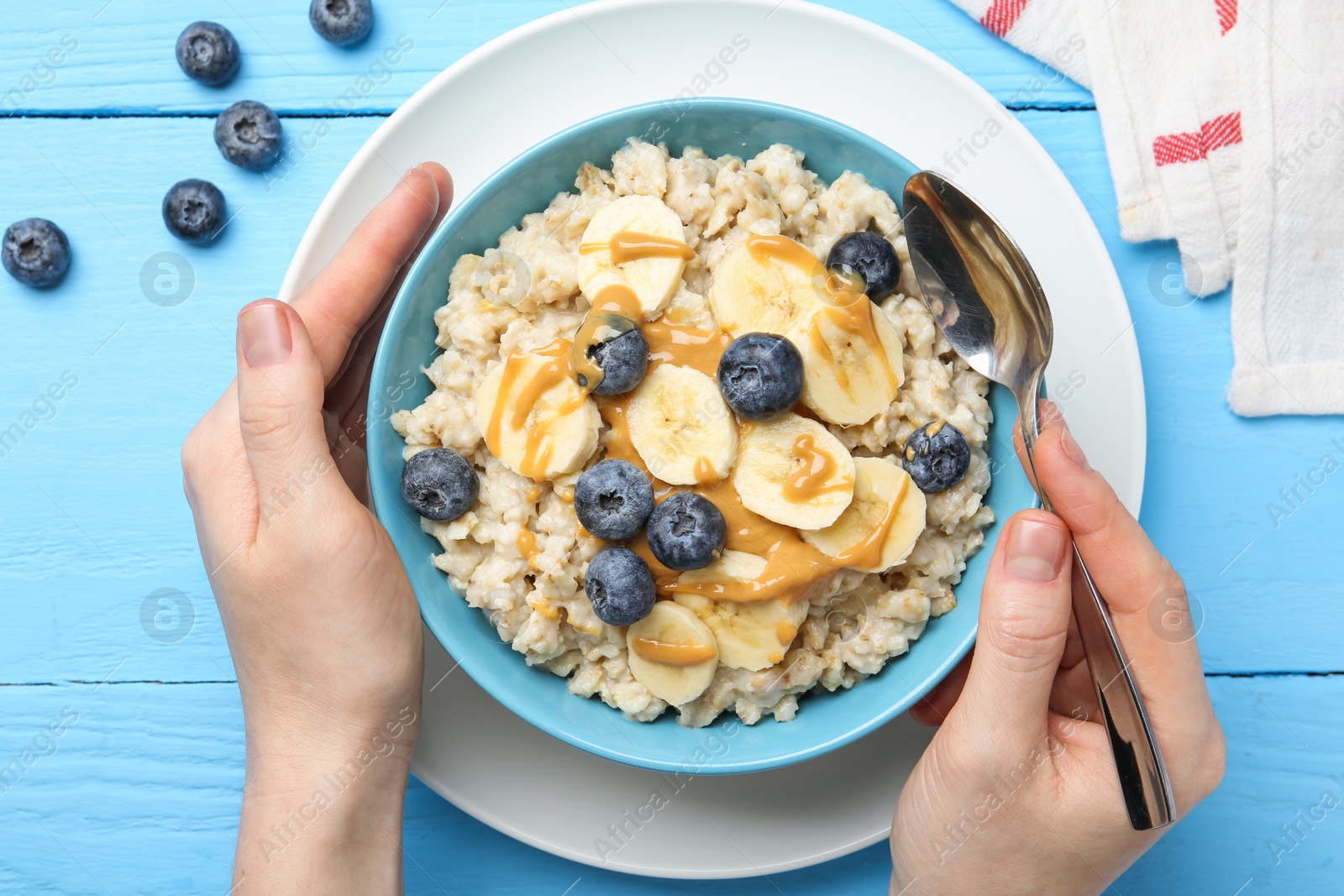 Photo of Woman eating tasty oatmeal with banana, blueberries and peanut butter at light blue wooden table, top view