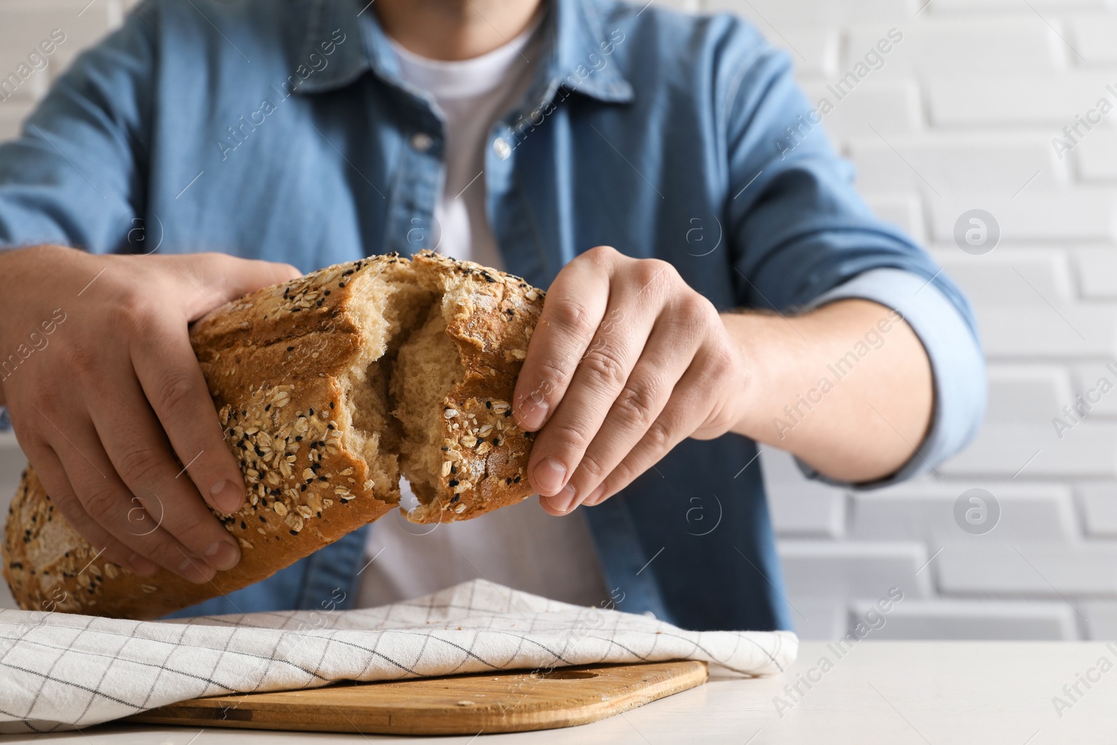 Photo of Man breaking loaf of fresh bread at white table near brick wall, closeup