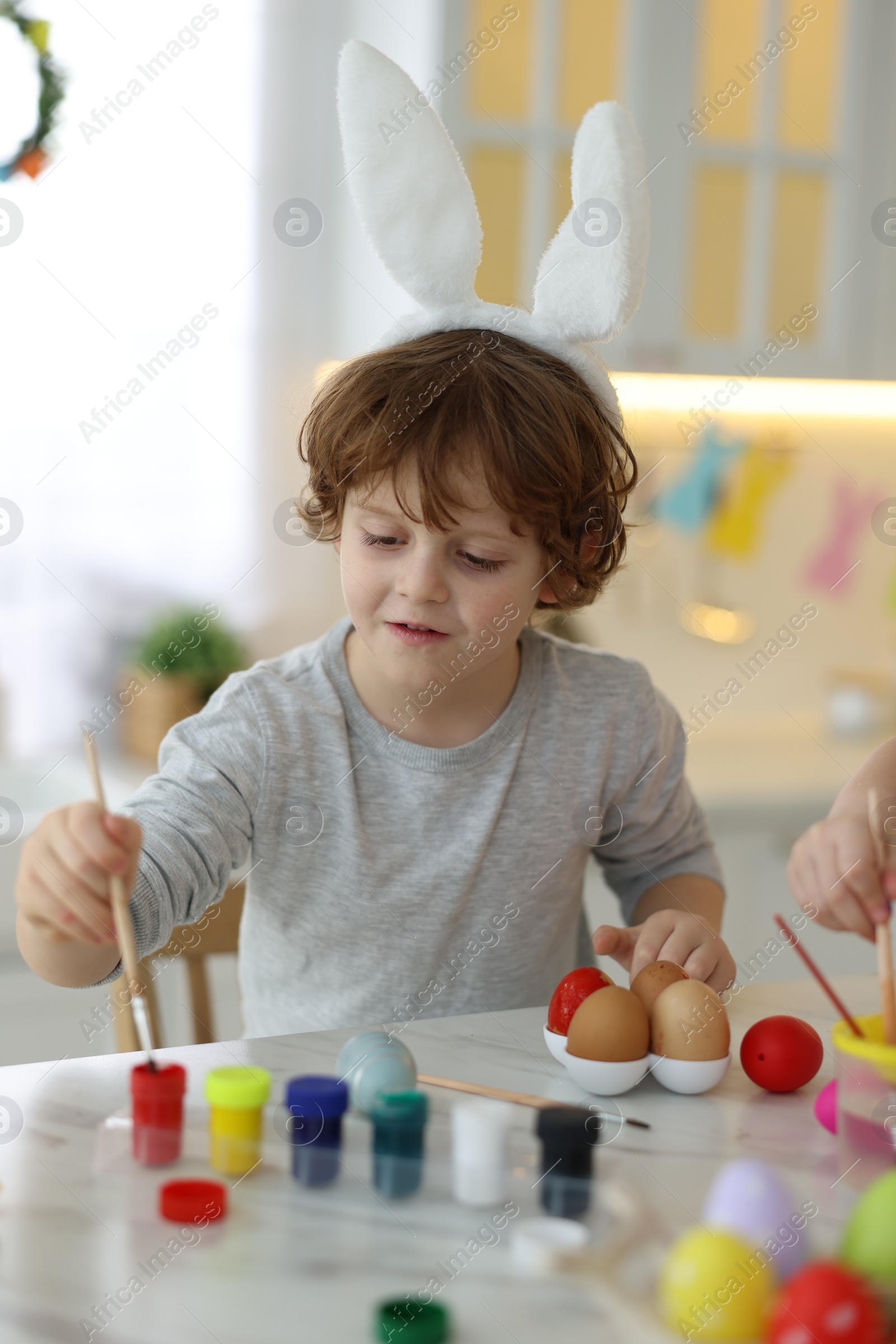 Photo of Easter celebration. Children with bunny ears painting eggs at white marble table in kitchen