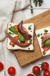 Delicious sandwiches with cream cheese, anchovies, tomatoes and basil on white wooden table, flat lay