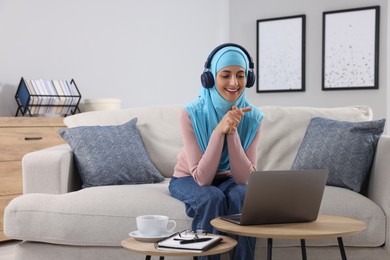 Muslim woman in headphones using laptop at wooden table in room. Space for text