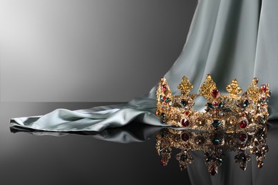 Photo of Beautiful golden crown with gems near light cloth on dark mirror surface. Space for text