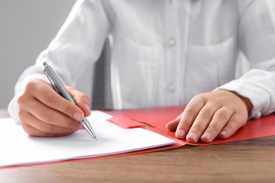 Photo of Woman writing on sheet of paper in red folder at wooden table in office, closeup