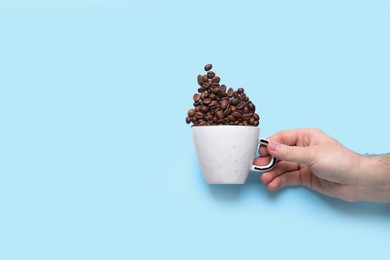 Man holding cup and scattered coffee beans on light blue background, top view. Space for text