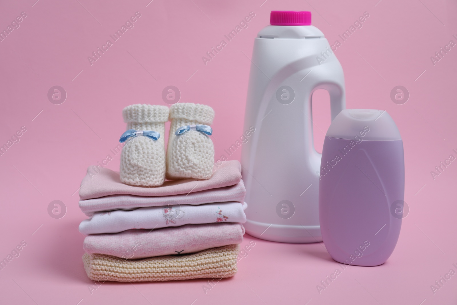 Photo of Laundry detergents, stack of baby clothes and small booties on pink background