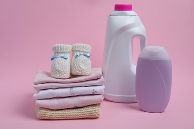 Laundry detergents, stack of baby clothes and small booties on pink background