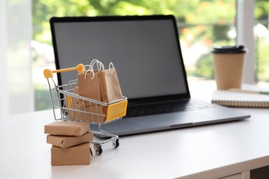 Photo of Internet shopping. Small cart with bags and boxes near laptop on table indoors, space for text