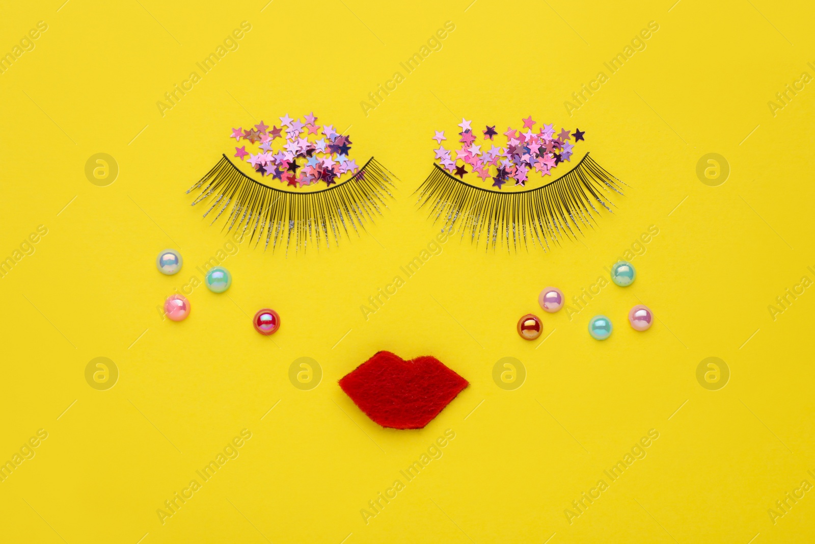 Photo of False eyelashes, red lips, beads and confetti as beautiful face on yellow background, flat lay