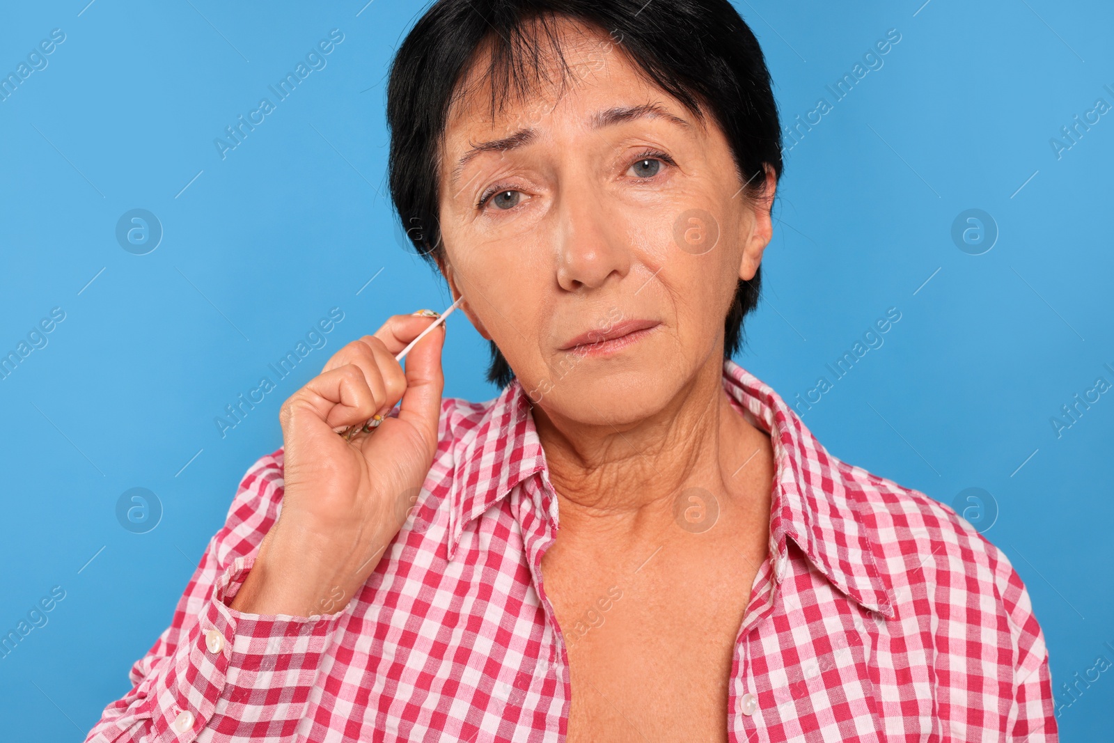 Photo of Senior woman cleaning ear with cotton swab on light blue background