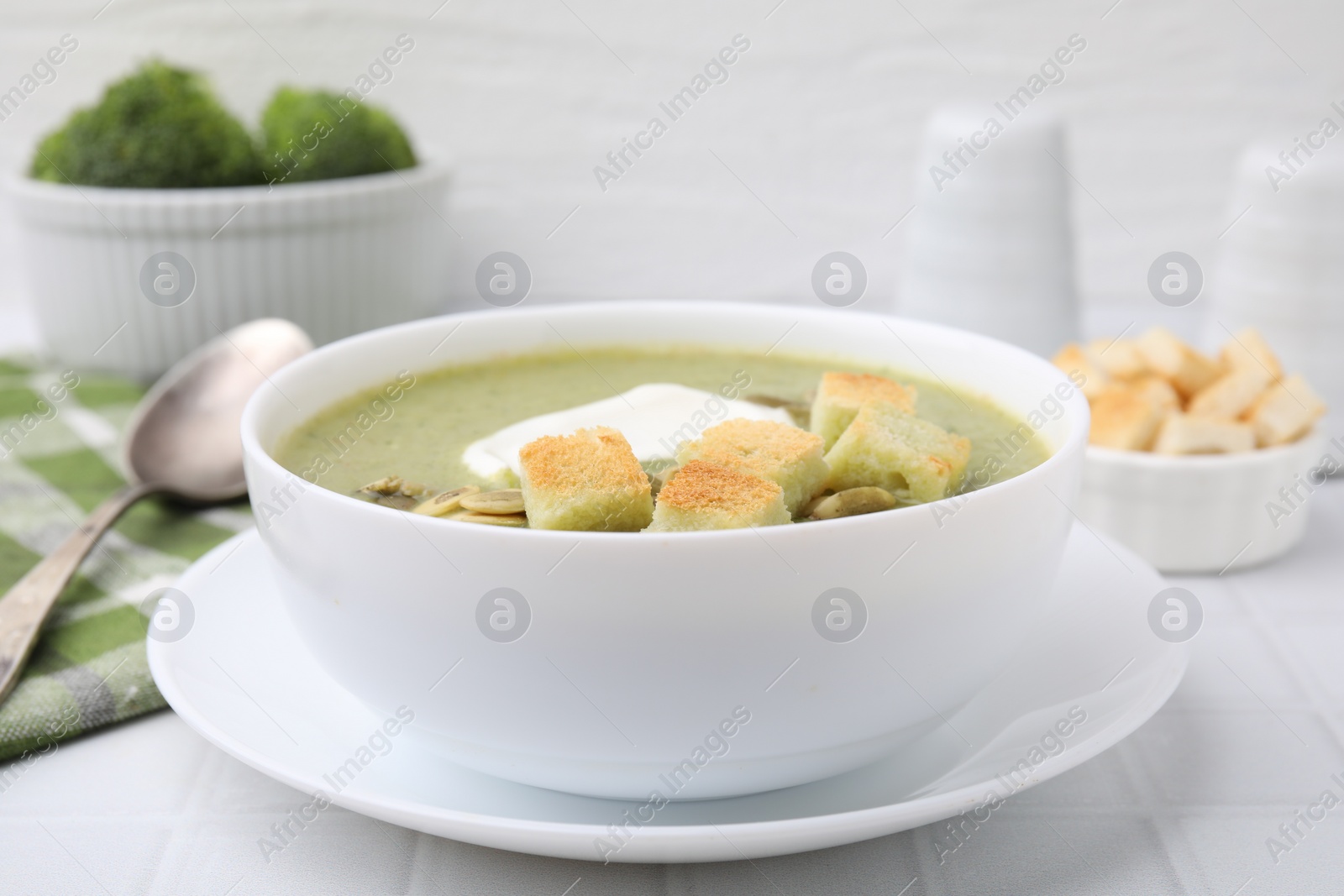 Photo of Delicious broccoli cream soup with croutons, sour cream and pumpkin seeds served on white tiled table, closeup