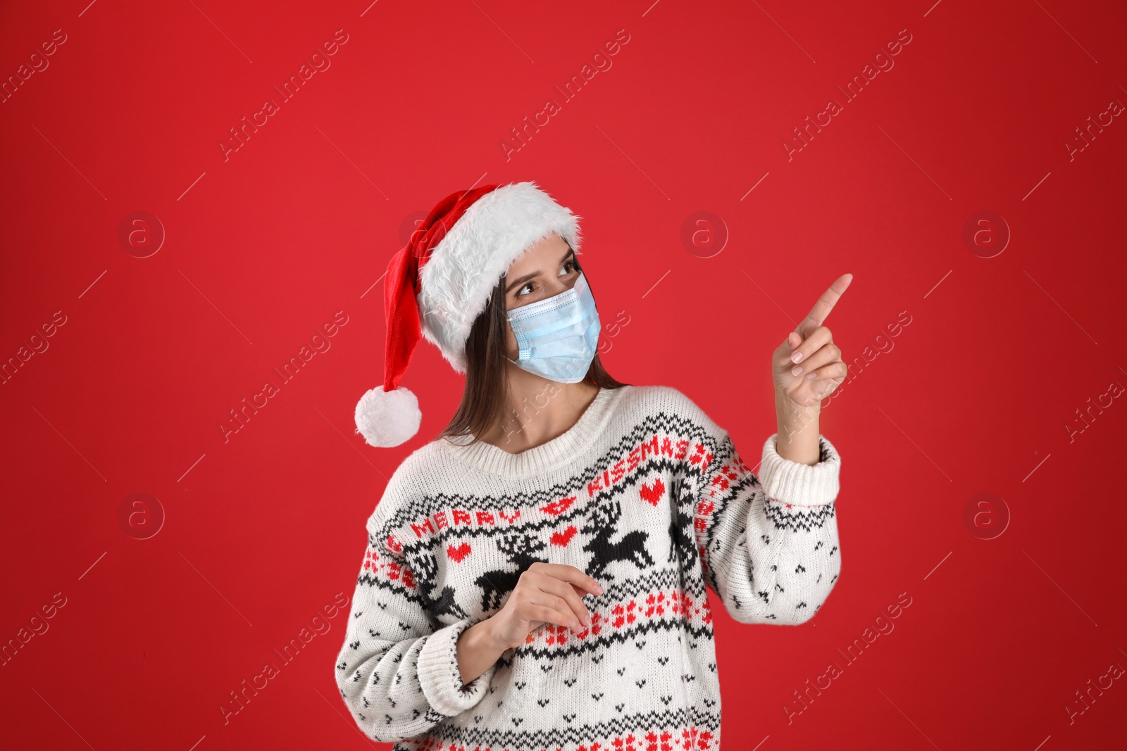 Photo of Pretty woman in Santa hat and medical mask pointing on red background