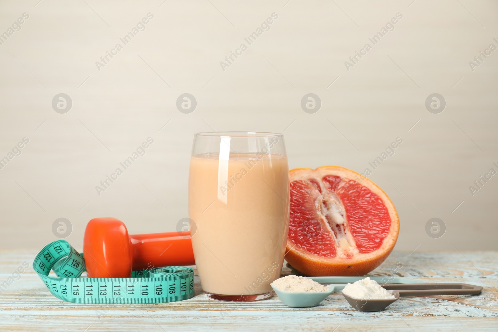 Photo of Tasty shake, grapefruit, dumbbell, scoops with different powders and measuring tape on wooden table against light background, space for text. Weight loss