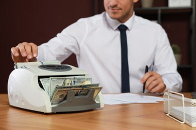Photo of Man putting money into banknote counter at wooden table indoors, closeup