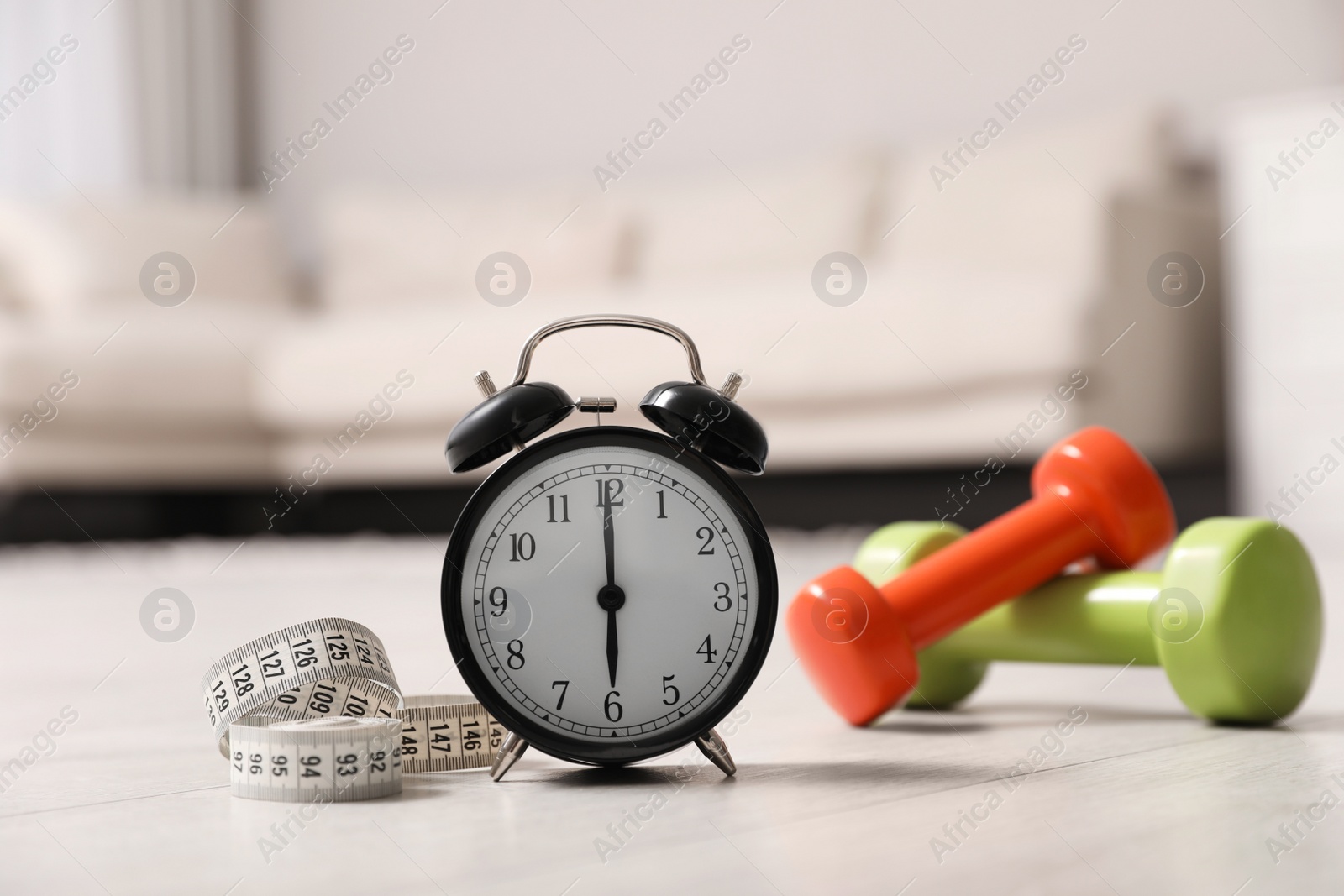 Photo of Alarm clock, measuring tape and dumbbells on wooden floor indoors. Morning exercise