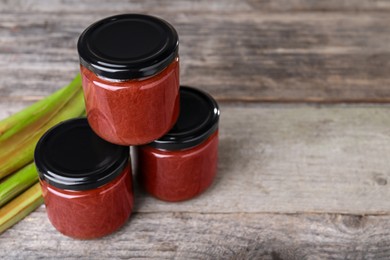 Photo of Jars of tasty rhubarb jam and stalks on wooden table, space for text