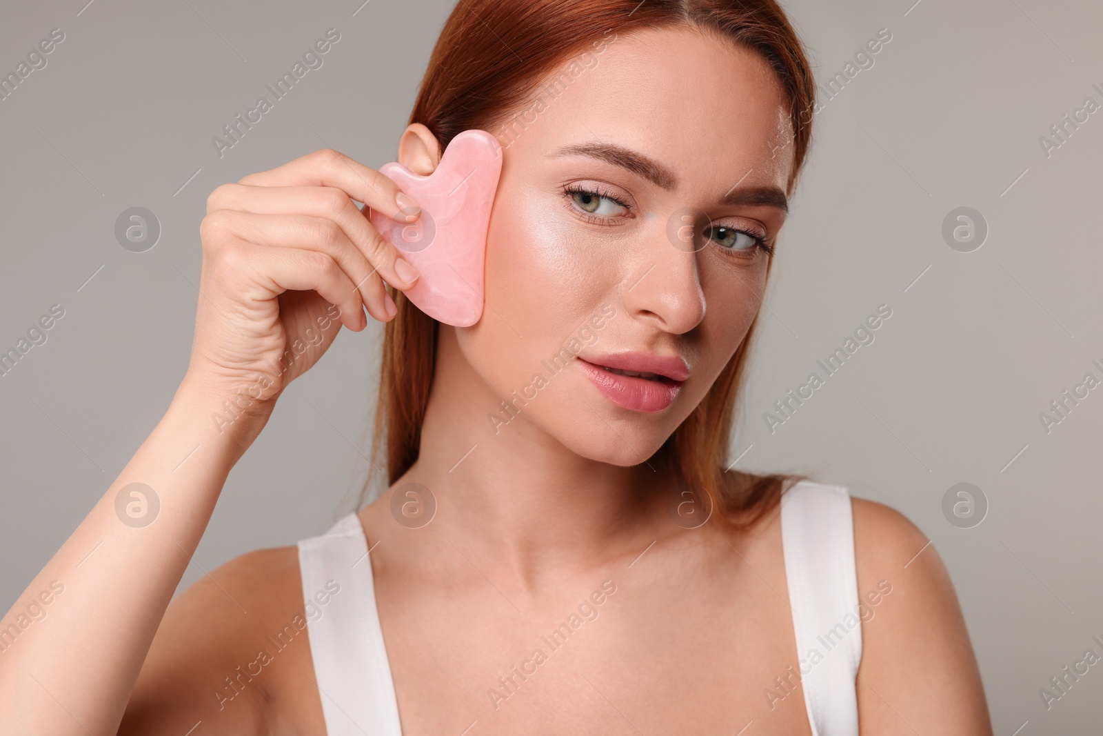 Photo of Young woman massaging her face with rose quartz gua sha tool on grey background