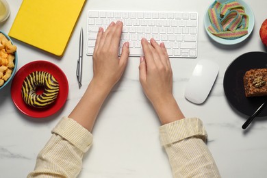 Photo of Bad eating habits. Woman working on computer at white marble table with different snacks, top view