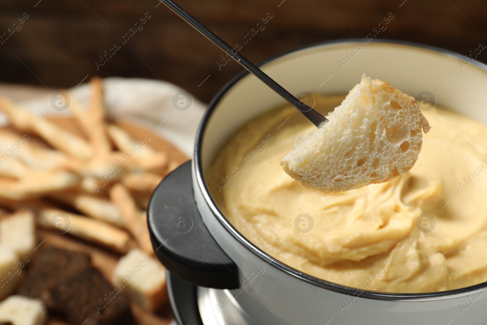 Photo of Dipping piece of bread into fondue pot with melted cheese on blurred background, closeup